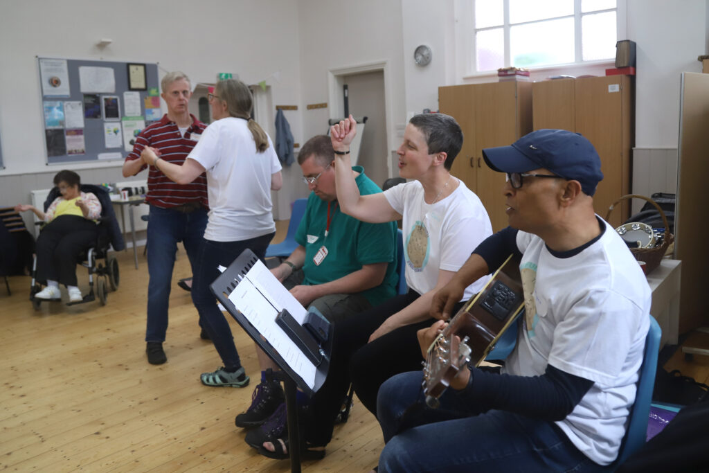 Doug is playing the guitar, Dawn is singing and Annette is dancing with a gentleman during a time of singing in the church hall.